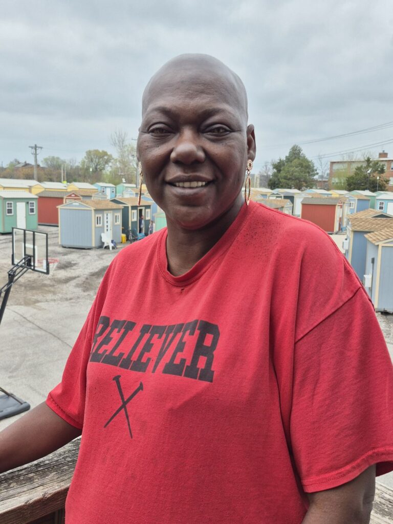A woman stands smiling on a balcony in front of a community of colorful tiny homes. She wears a red shirt that reads “BELIEVER” with two crossed nails beneath the text.