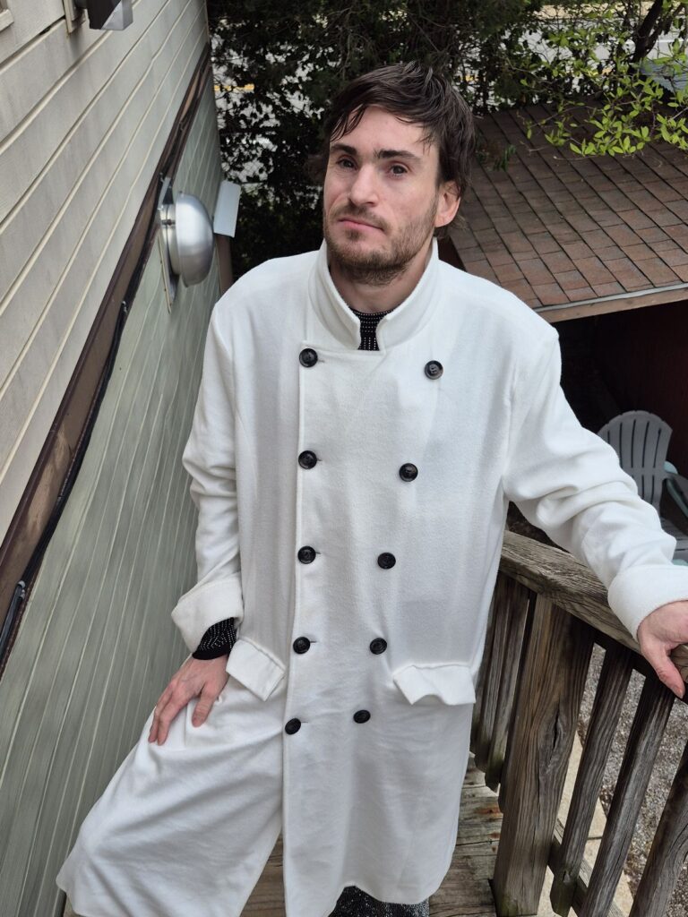 A man stands confidently on an outdoor wooden staircase, wearing a long white coat with black buttons. He looks directly at the camera with a calm expression.