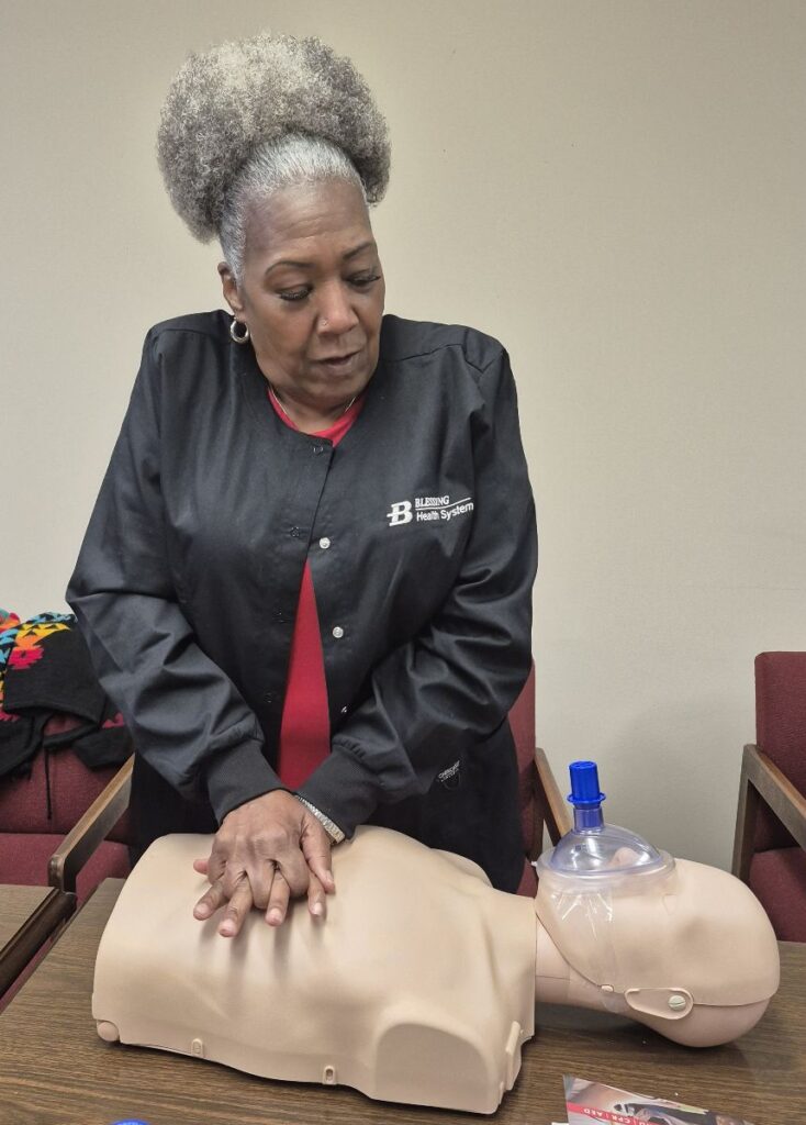 A woman in a black “Blessing Health System” jacket performs CPR compressions on a training mannequin during a certification session.
