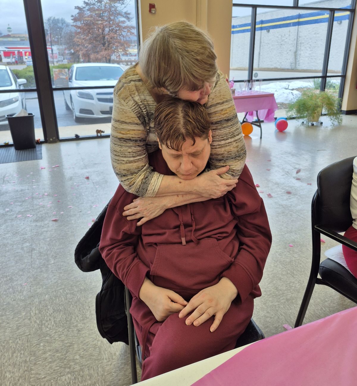 A client gently hugs another seated client from behind, resting her head on hers as she sits with his eyes closed. They are in a bright room with large windows and pink decorations in the background.