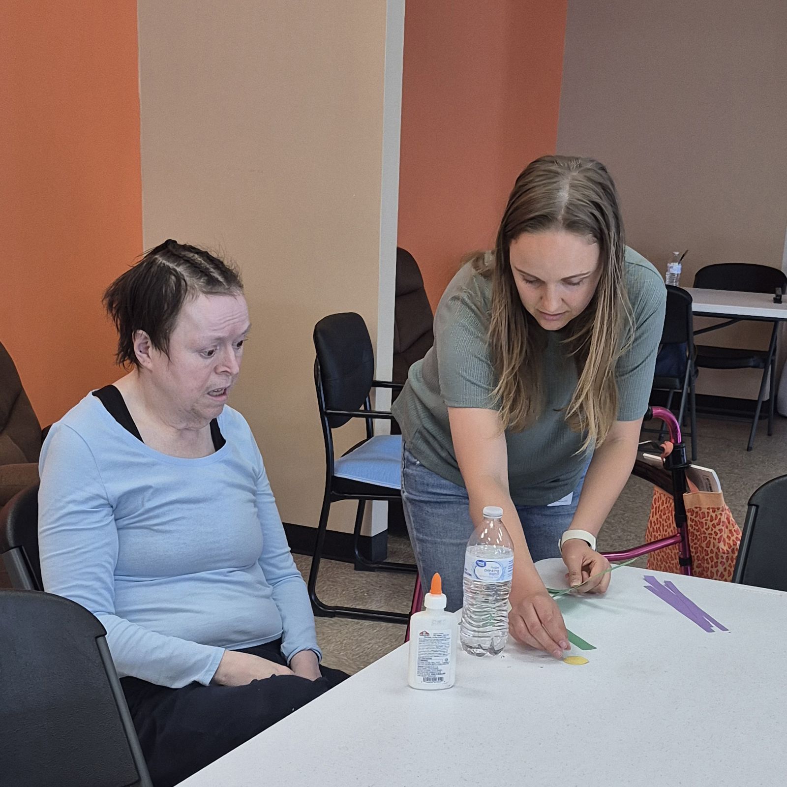 A Occupational Therapist at Magdala House helps a client with a craft project using colorful paper strips at a table. Glue and a water bottle are on the table.