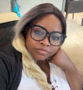 Portrait of Carmen Woodson, a Magdala House staff member at Tiny Homes St. Louis, wearing black glasses and a blonde braid while sitting thoughtfully with a calm expression.