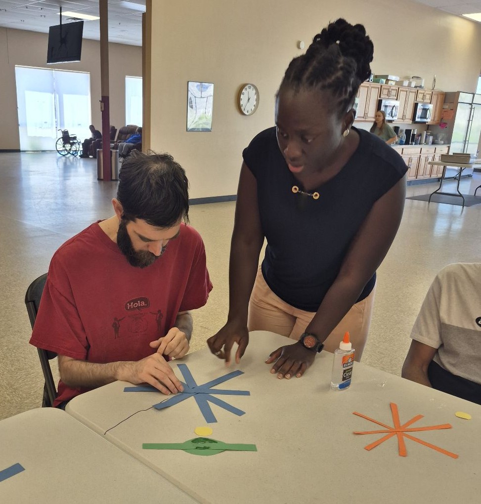 A woman stands beside a seated man as he carefully assembles a paper flower using construction paper and glue during an occupational therapy session at Magdala House’s Adult Day Care Program.