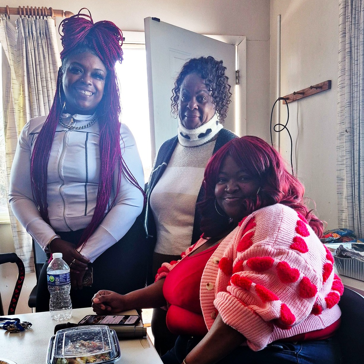 Three women at Magdala House Tiny Homes smile together during a Women’s Group gathering in St. Louis. One woman is seated at a table while two others stand beside her in a warm, welcoming community space.